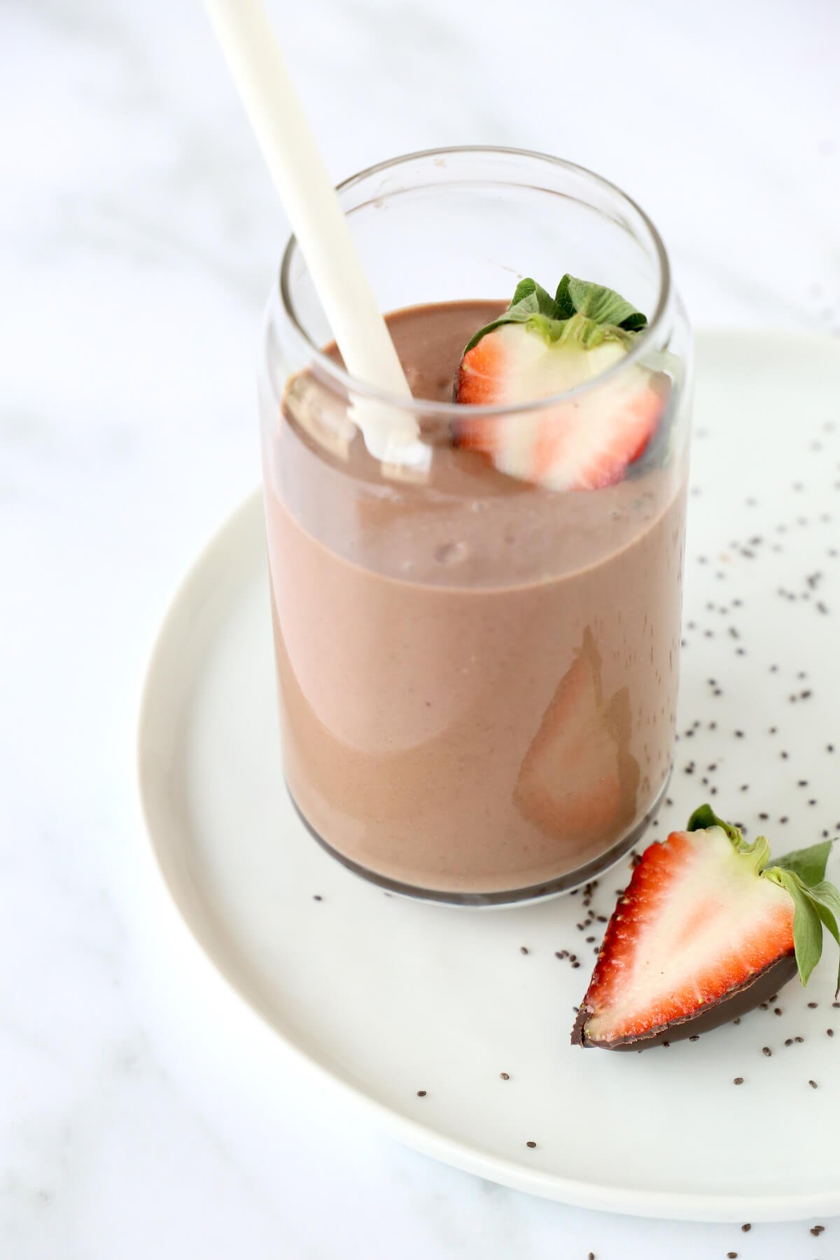 A clear glass sitting on a white plate filled with chocolate brown smoothie, a white straw and a sliced strawberry.  
