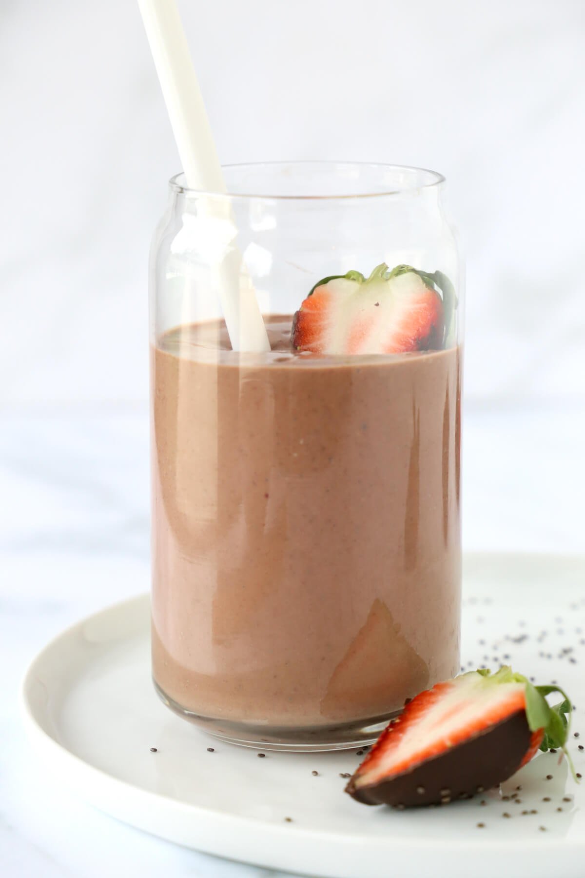 A clear glass sitting on a white plate filled with chocolate brown smoothie, a white straw and a sliced strawberry.
