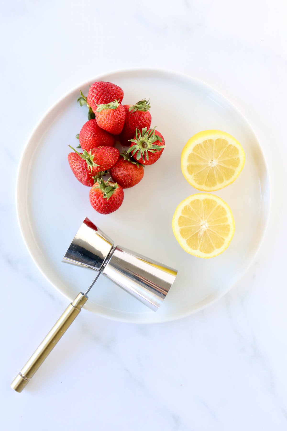 A white plate with whole strawberries, a lemon cut in half and a cocktail measuring spoon.