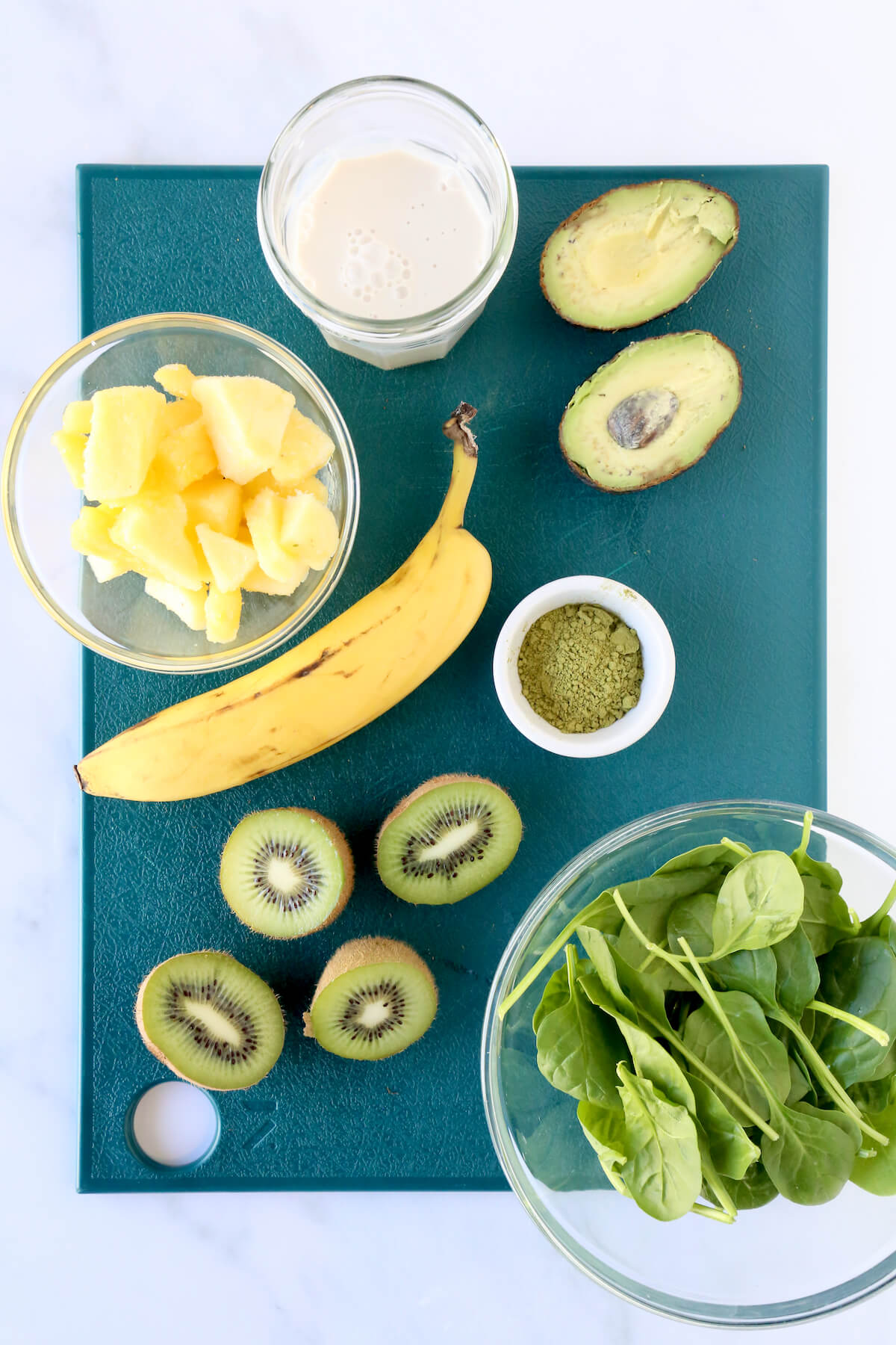 A green cutting board with almond milk, sliced avocado, frozen pineapple, one banana, sliced kiwi, a bowl of matcha powder and a bowl of spinach.