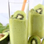 Two clear glasses filled with green smoothie and a straw with sliced kiwi.