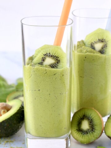 Two clear glasses filled with green smoothie and a straw with sliced kiwi.