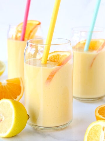 Three clear glasses with orange smoothie, a pink straw, yellow straw and blue straw.