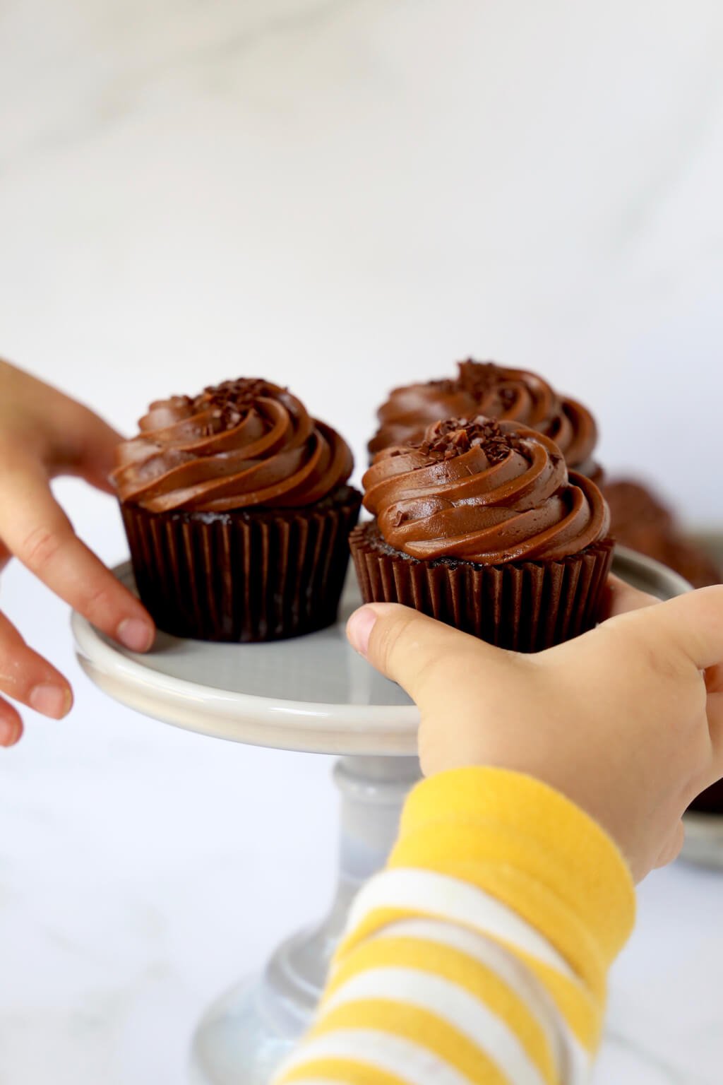 A gray cake stand holding three cupcakes with two hands reaching for the cupcakes.  