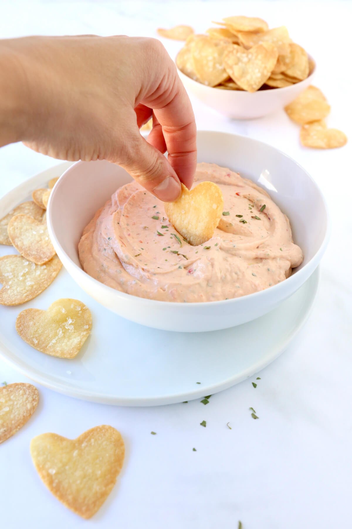 A white bowl with pink dip and a hand dipping a heart chip.  