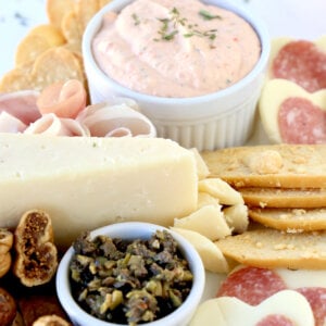 A white square platter filled with heart tortilla chips, prosciutto, cheese, heart salami and dried figs.