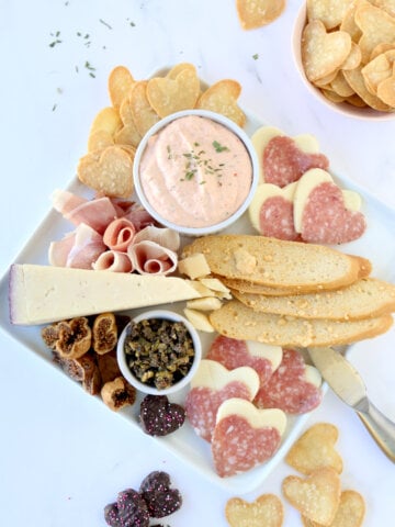 A white square platter filled with heart tortilla chips, prosciutto, cheese, heart salami and dried figs.
