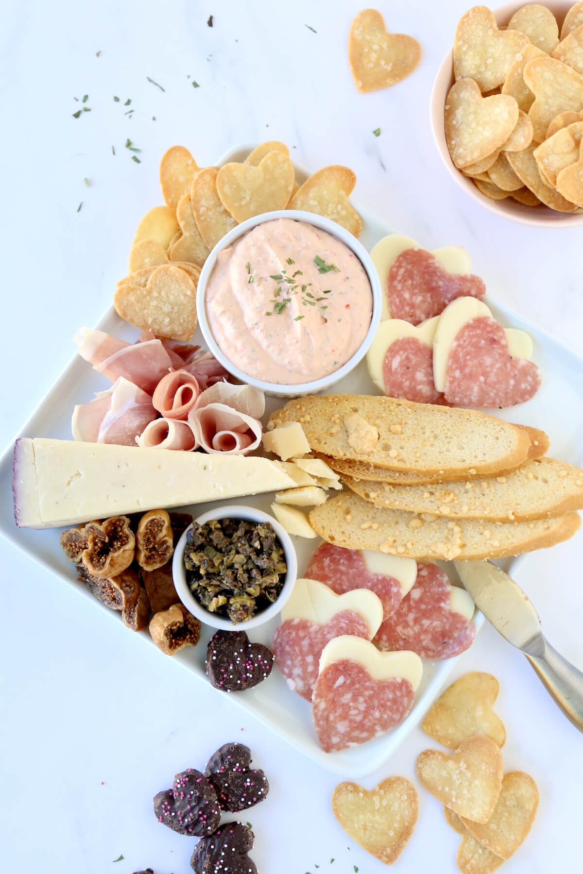 A white square platter filled with heart tortilla chips, prosciutto, cheese, heart salami and dried figs.  