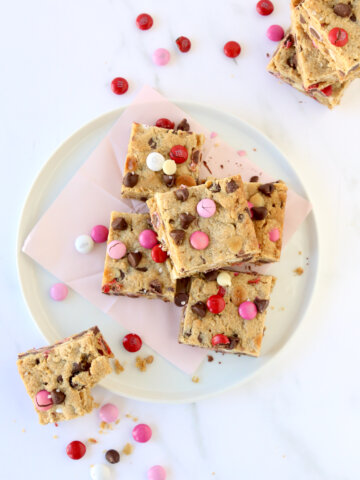Cookie bar squares stacked on top of each other on a white plate with pink, red and white m&m candies.