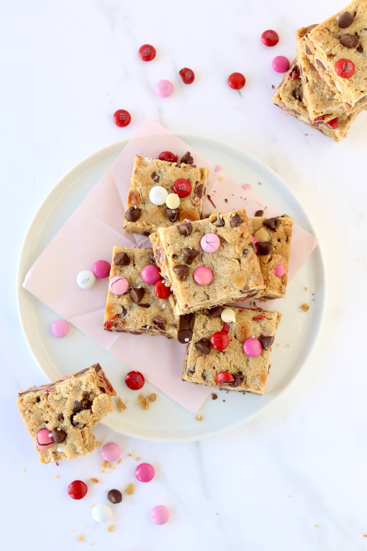 Cookie bar squares stacked on top of each other on a white plate with pink, red and white m&m candies.  