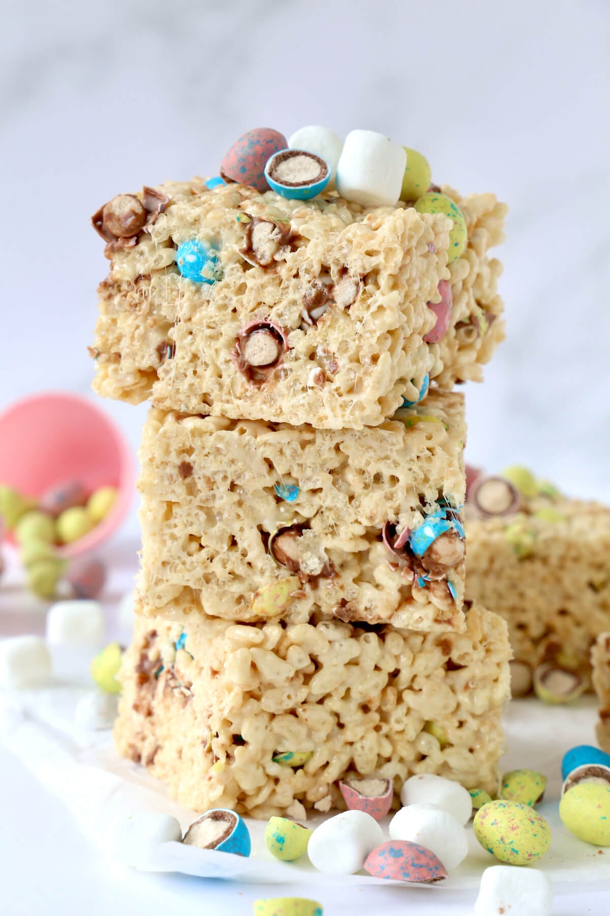 Three square rice krispie cereal treats are stacked on teacher other with the malted egg candies and marshmallows sprinkled around. 