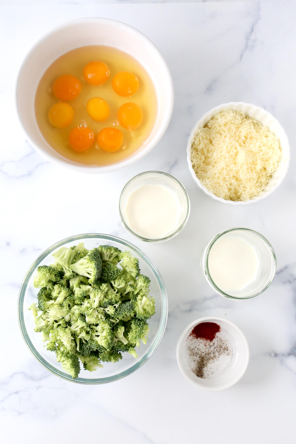 A bowl of eggs, shredded cheese, broccoli, a jar of milk and heavy cream and a small bowl of salt, pepper and paprika.