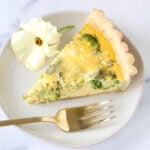 A slice of quiche on a plate with a gold fork and a white flower.
