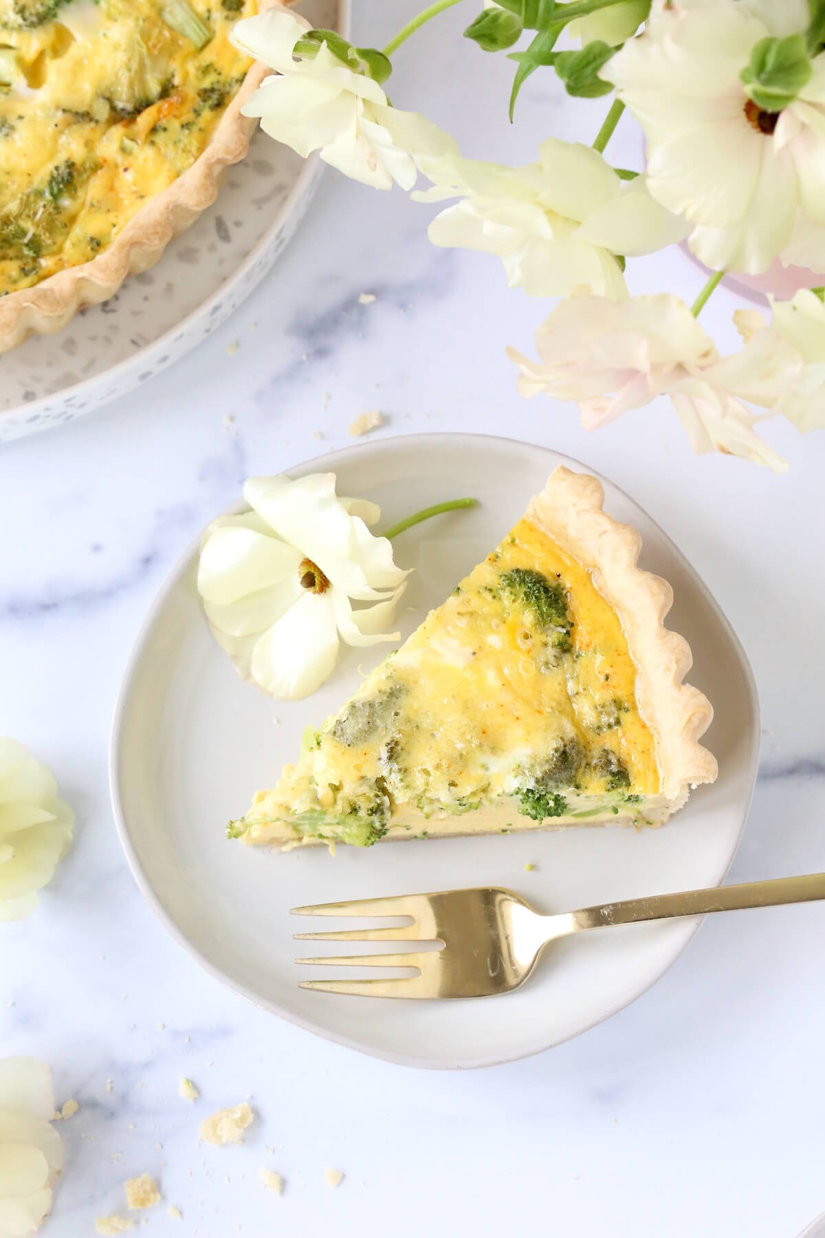 A slice of quiche on a plate with a gold fork and a white flower.