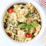 A white bowl filled with orzo, shrimp, tomatoes next to a piece of french bread and a stack of plates.