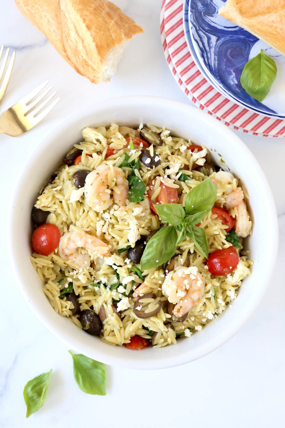 A white bowl filled with orzo, shrimp, tomatoes next to a piece of french bread and a stack of plates.  