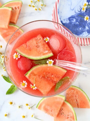 A punch bowl filled with a red drink, sliced watermelons, watermelon ice cubes and fresh flowers.