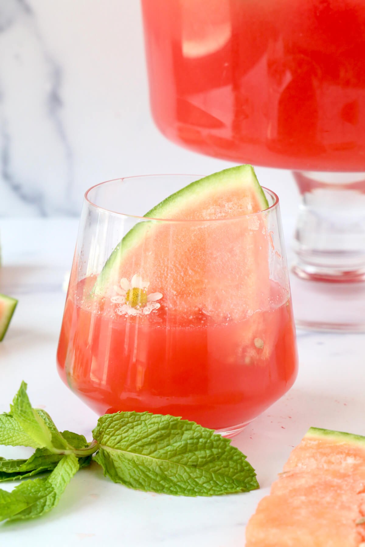 A glass filled with a red drink and a slice of watermelon and one white flower.  
