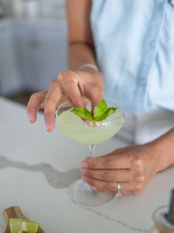 A glass with a light green liquid and a person putting a piece of fresh mint in the glass.