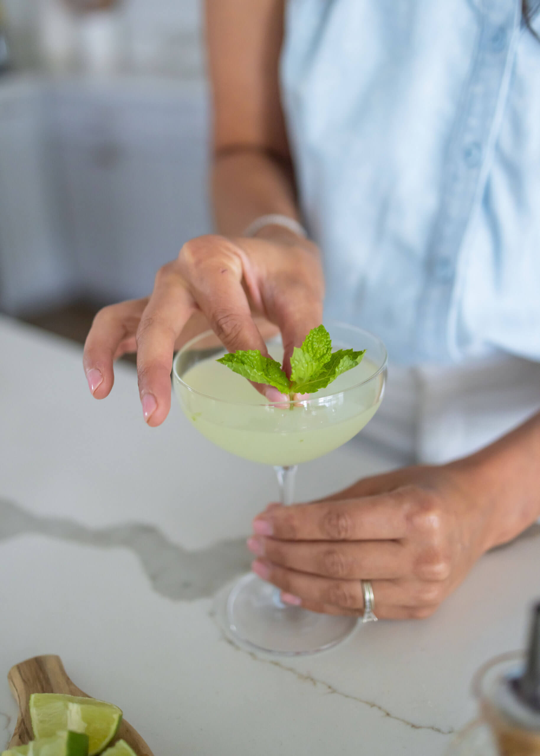 A glass with a light green liquid and a person putting a piece of fresh mint in the glass.  