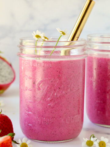 A clear glass with bright pink smoothie inside and a gold straw and small white flowers.