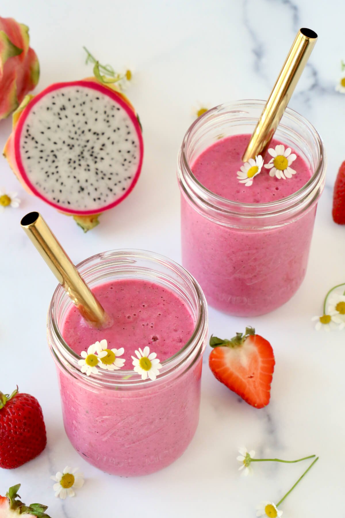A clear glass with bright pink smoothie inside and a gold straw and small white flowers.