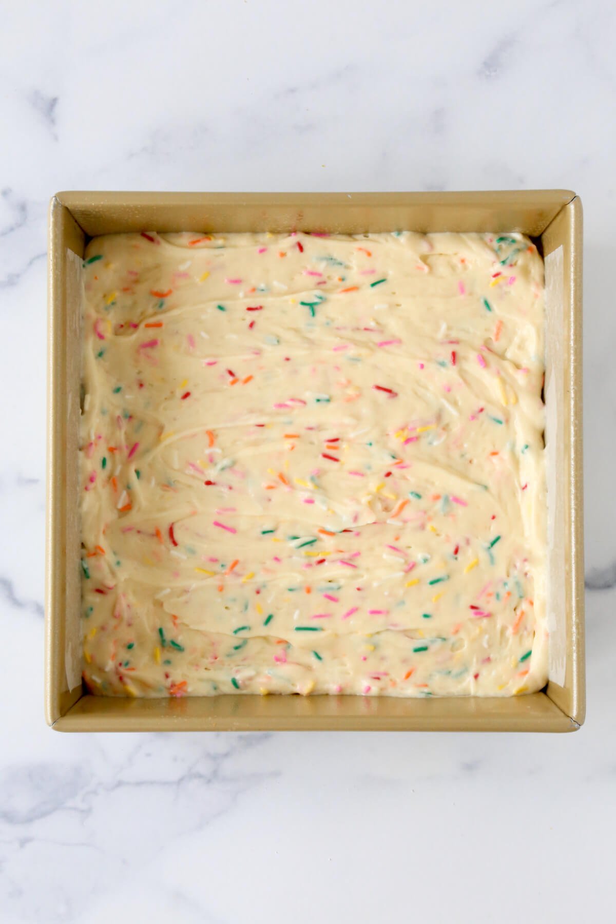 A square gold cake pan with white cake batter with rainbow sprinkles.