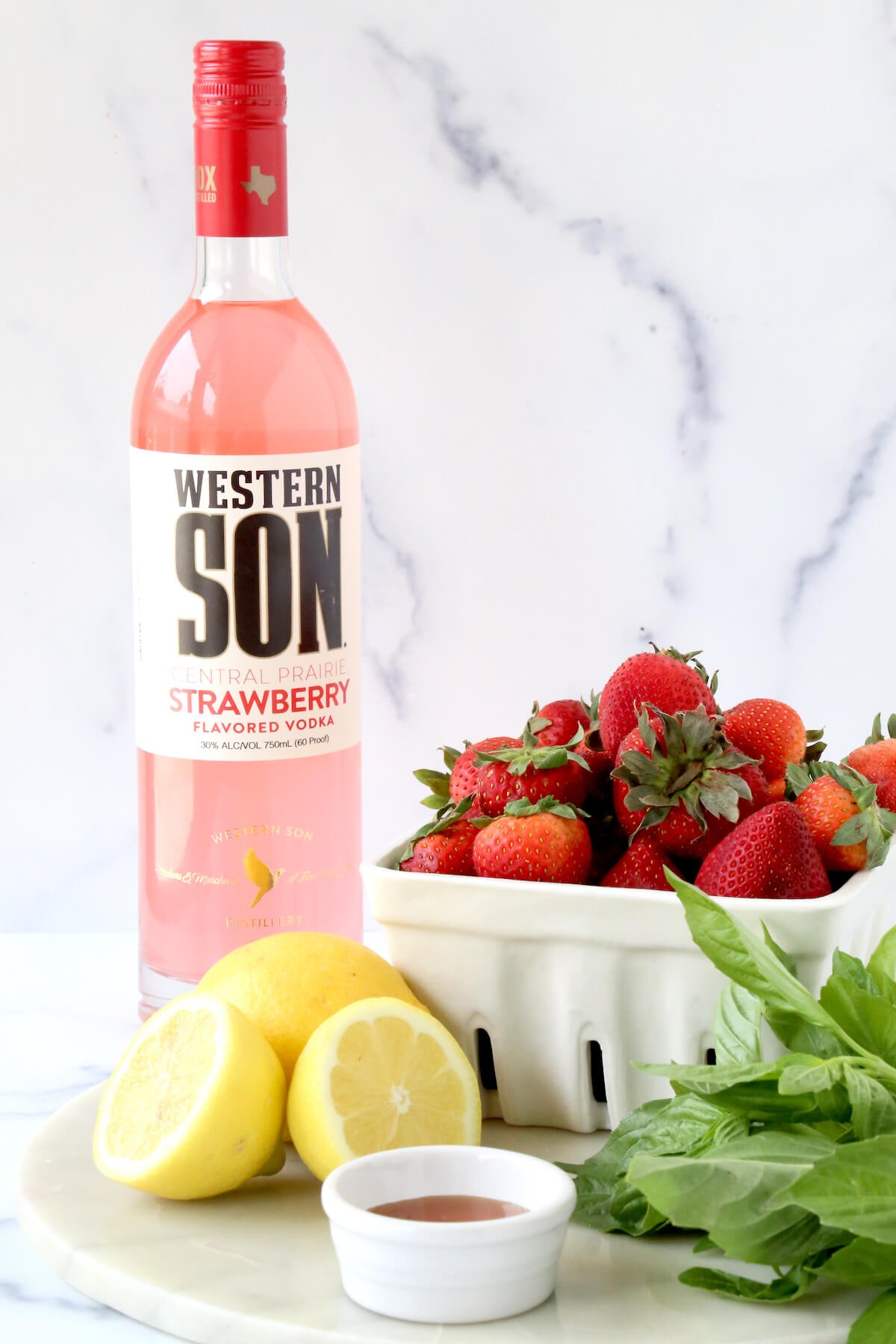 A tall bottle filled with a pink liquid next to sliced lemons and a bowl of fresh strawberries.