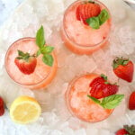 A glass filled with a red liquid and a fresh strawberry and a mint leaf surrounded by ice and a lemon slice.