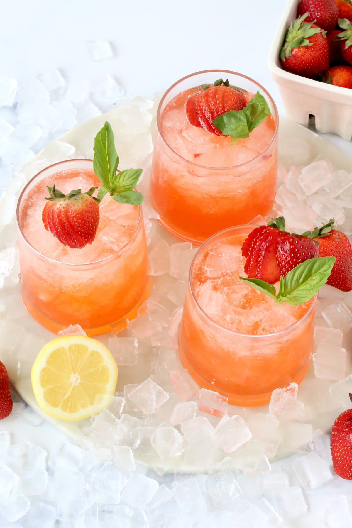 A glass filled with a red liquid and a fresh strawberry and a mint leaf surrounded by ice and a lemon slice.  