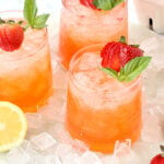 A glass filled with a red liquid and a fresh strawberry and a mint leaf surrounded by ice and a lemon slice.