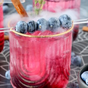 A glass filled with a pink drink with blueberries on top and smoke coming out.