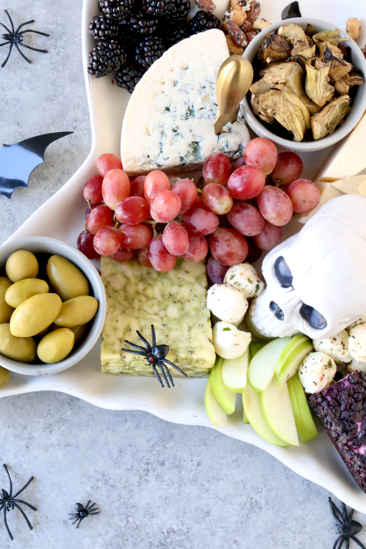 A ghost platter filled with blackberries, grapes, apple slices, three cheese wedges, olives and artichoke hearts.