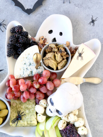 A ghost platter filled with blackberries, grapes, apple slices, three cheese wedges, olives and artichoke hearts.