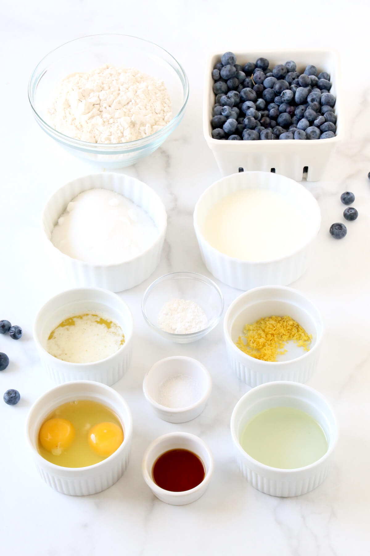 White bowls filled with flour, blueberries, sugar, eggs, vanilla extract,  vegetable oil and melted butter. 