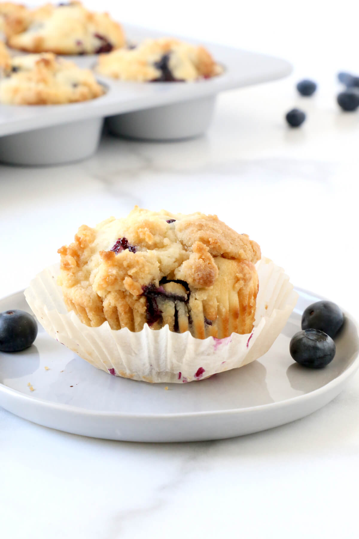 A gray plate with a muffin on top with the wrapper peeled off and blueberries next to it. 