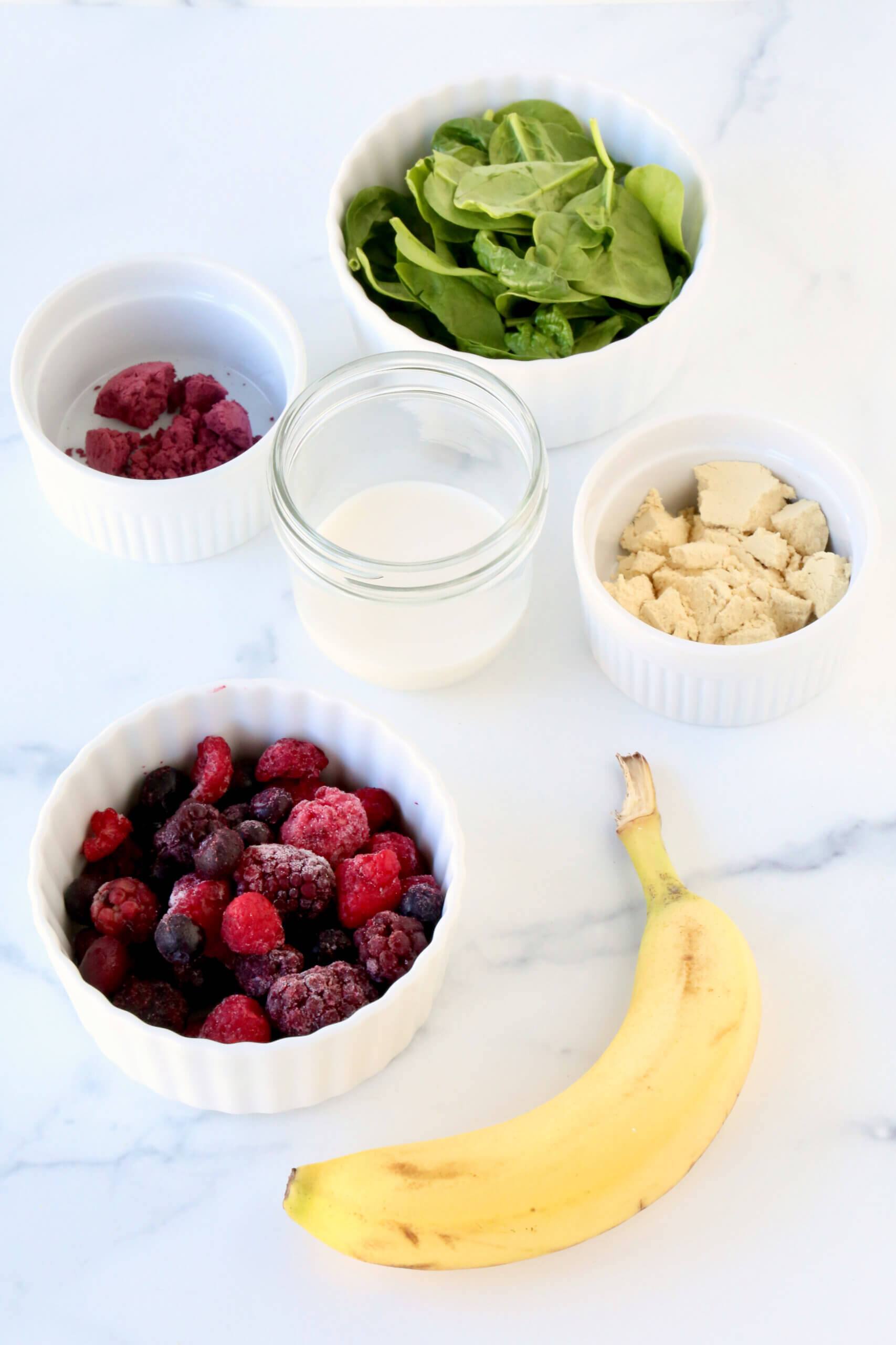 Bowls filled with spinach, mixed berries, protein powder, açaí powder, a jar with almond milk and a banana.  