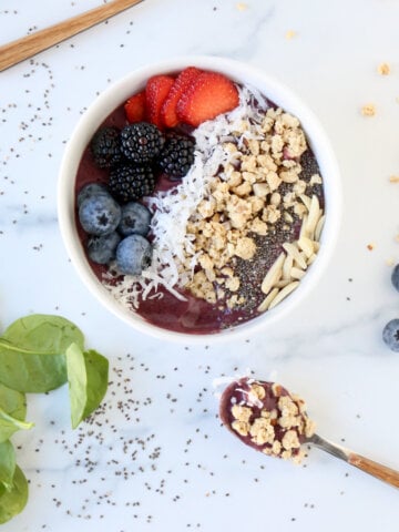 A white bowl filled with a purple smoothie and topped with granola, strawberries, blackberries, blueberries, coconut and almonds.