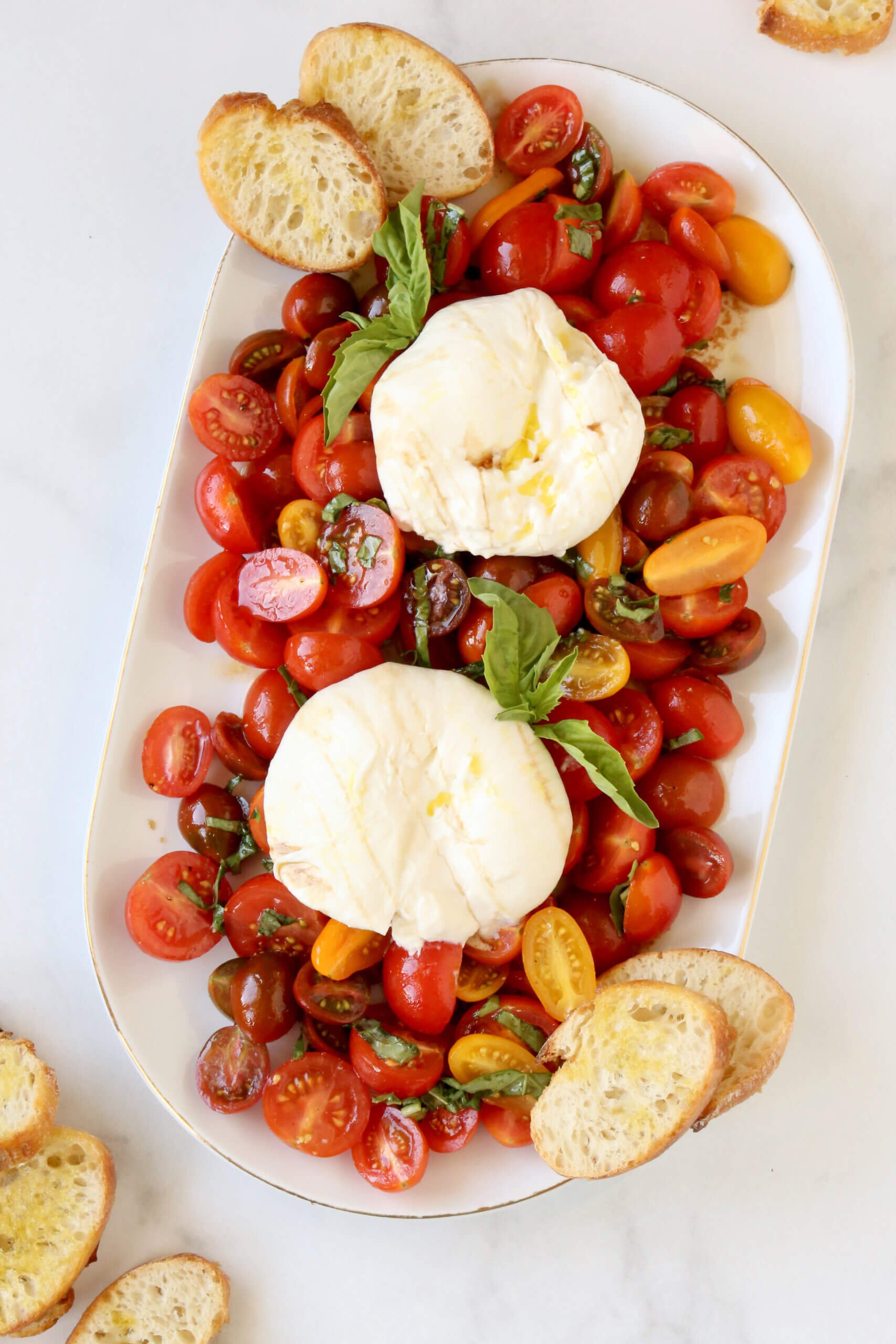 A white oval dish filled with sliced tomatoes, white cheese balls, basil and slices of toasted bread.  