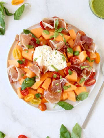 A white plate filled with orange melon, red, yellow and orange tomatoes, white cheese in the middle and green basil leaves.