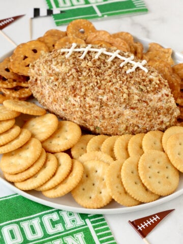 A white plate filled with crackers, pretzels and a football shaped cheese ball.