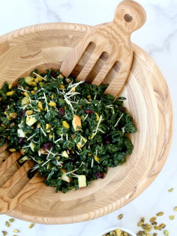 A wood bowl filled with green kale, pistachios, white pieces of cheese and small square apple slices.