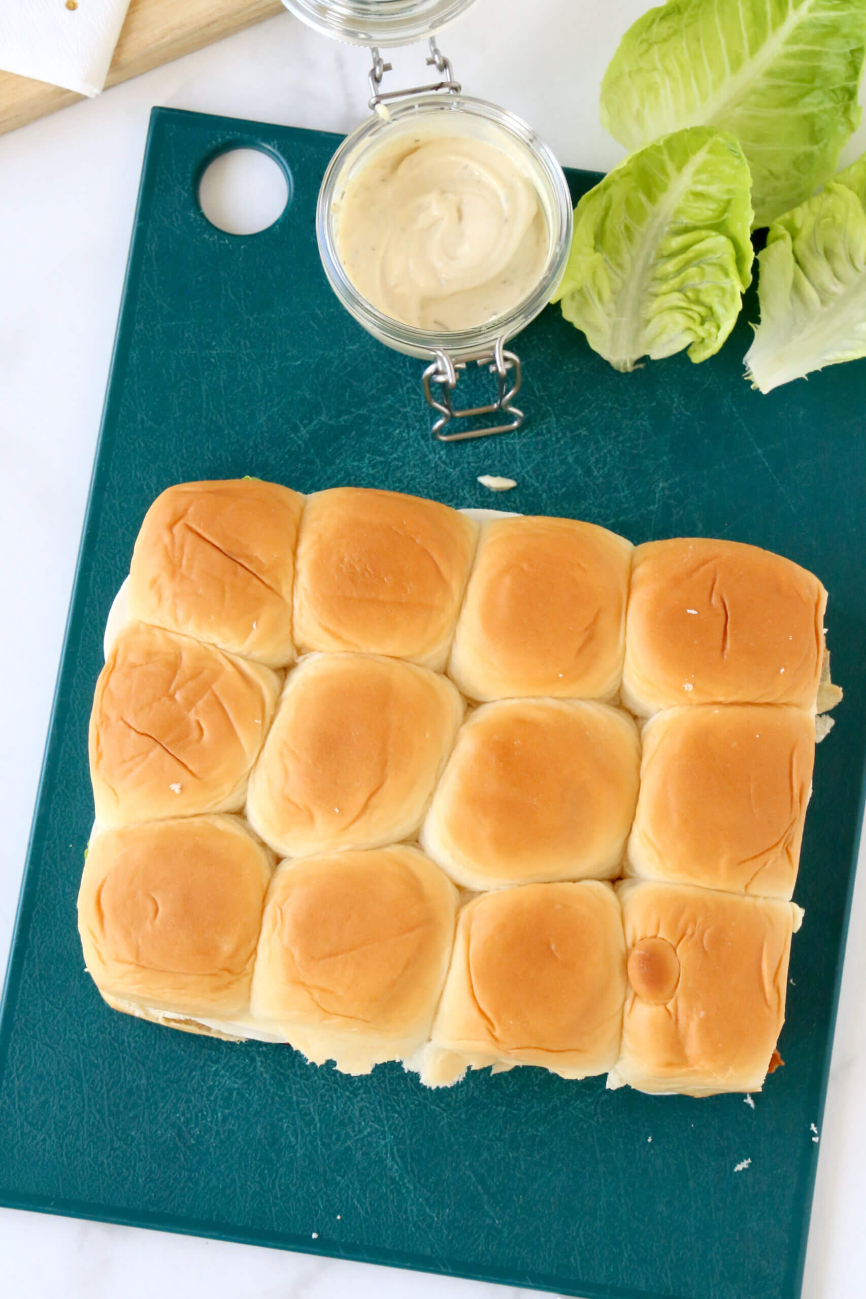 A green cutting board with bread rolls sitting on top with a jar of mayo dijon spread and lettuce leaves.  