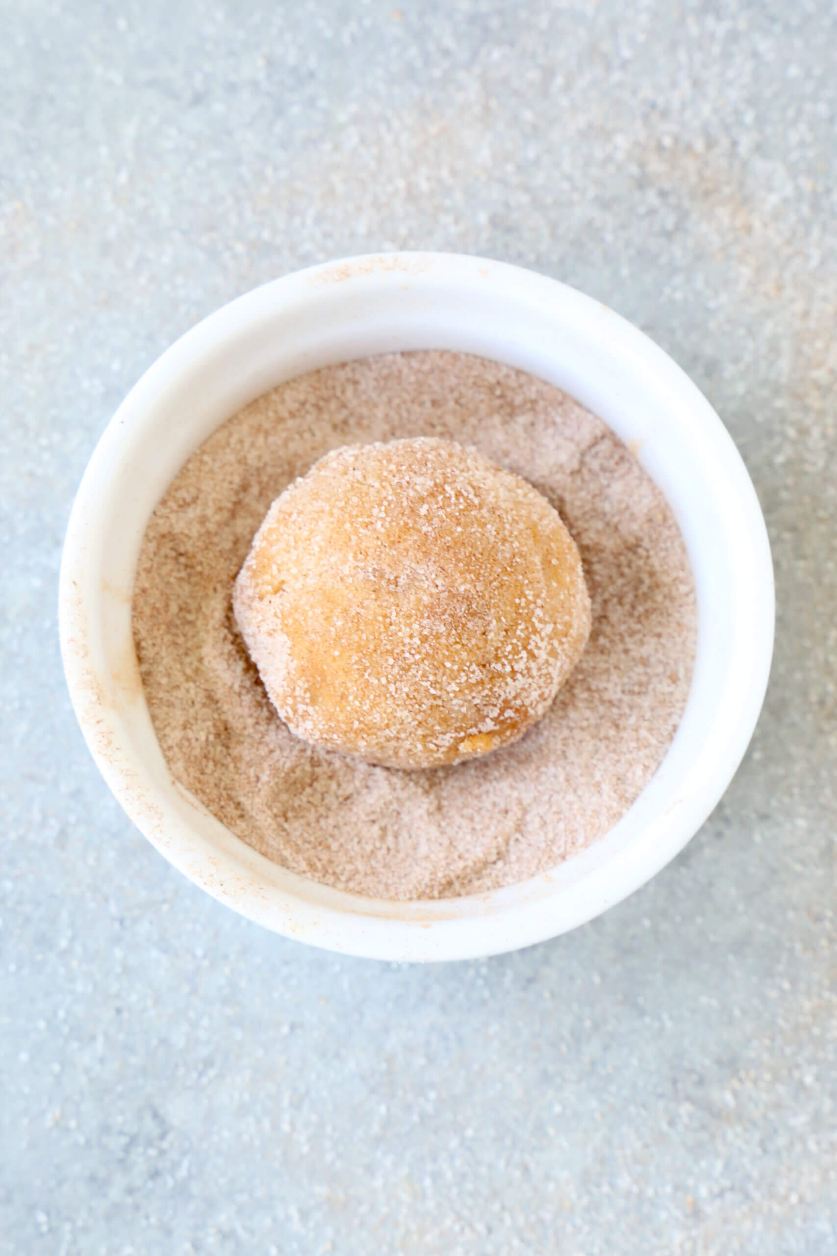 A white bowl filled with light brown cinnamon sugar mixture and one round cookie dough ball on top. 