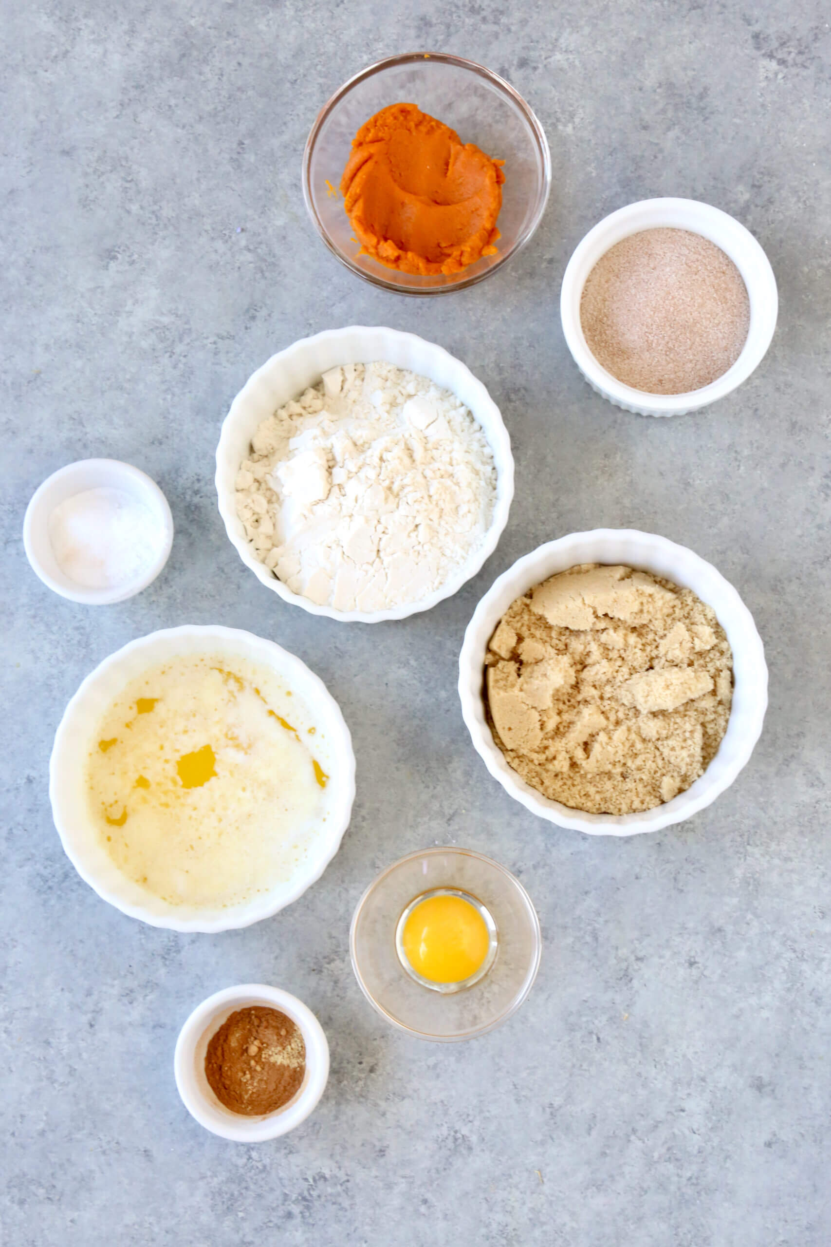 Eight bowls sitting on a gray surface filled with pumpkin puree, flour, brown sugar, melted butter, egg yolk, cinnamon sugar and pumpkin spice seasoning.