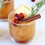 Three glasses filled with a light brown liquid, cranberries, an apple slice and a cinnamon stick.