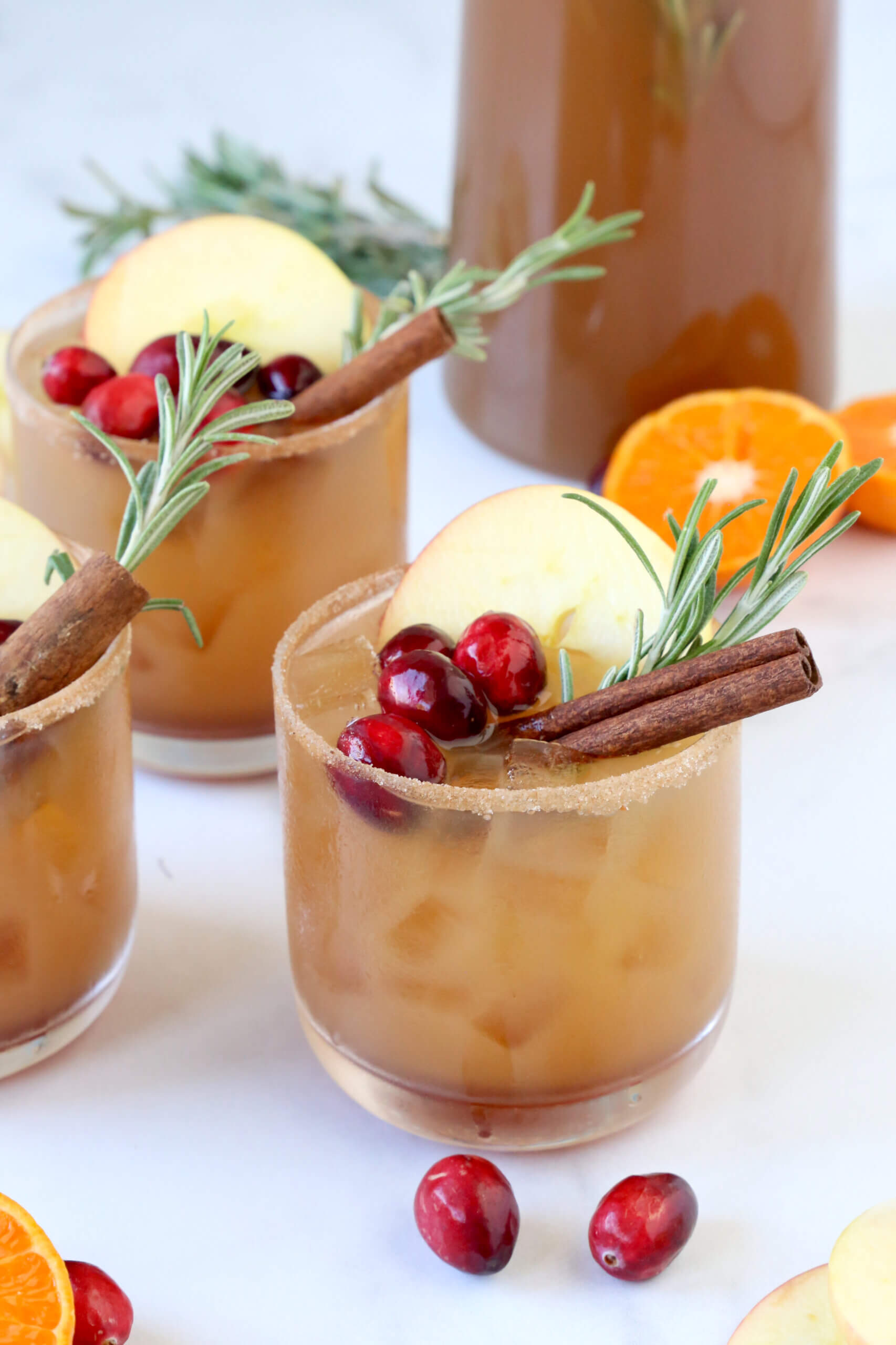 Three glasses filled with a light brown liquid, cranberries, an apple slice and a cinnamon stick.  