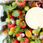 A round white platter topped with fresh strawberries, blackberries, green grapes, orange dried apricots and green fresh mint.