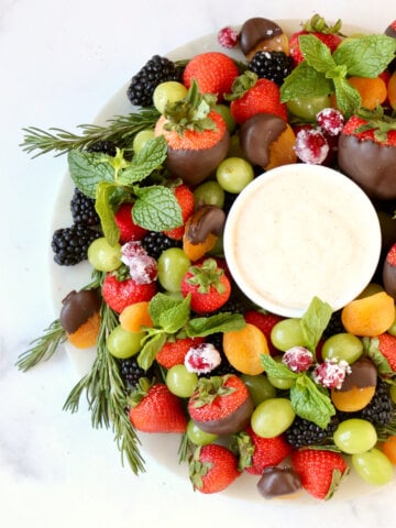 A round white platter topped with fresh strawberries, blackberries, green grapes, orange dried apricots and green fresh mint.