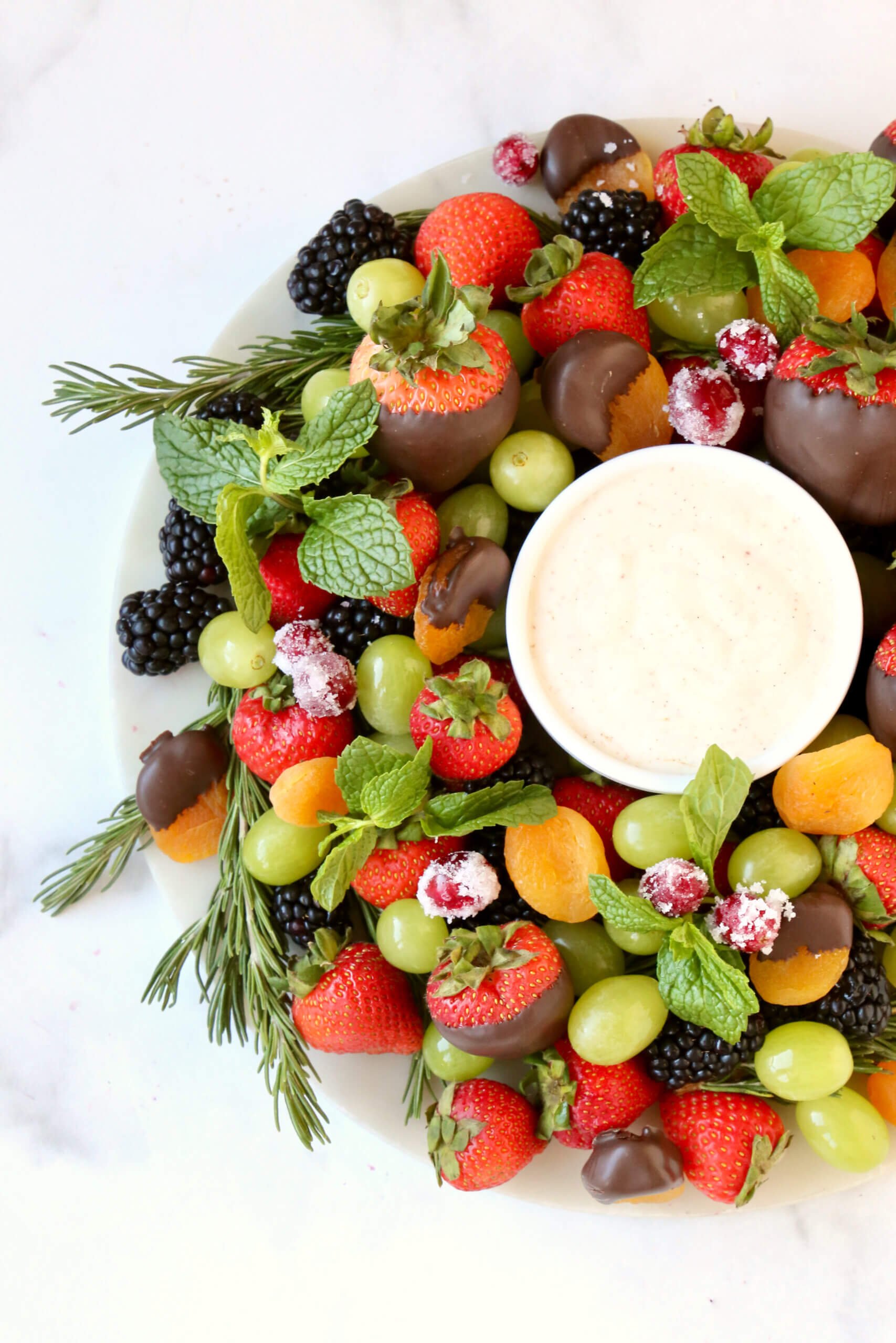 A round white platter topped with fresh strawberries, blackberries, green grapes, orange dried apricots and green fresh mint.